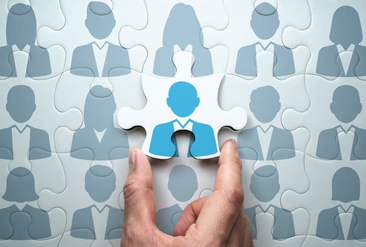 A puzzle made up of the grey silhouettes of businesspeople. A hand is placing the final puzzle piece, which has a blue silhouette