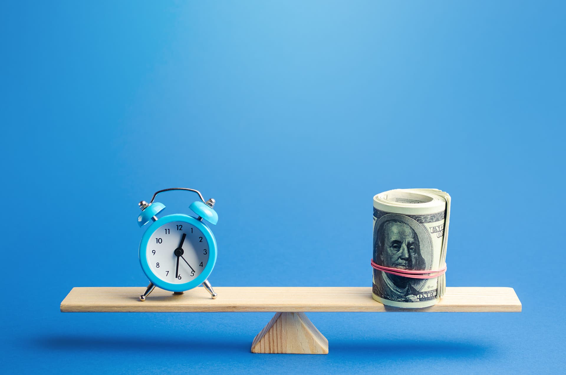 Blue alarm clock and a bundle of dollars balancing on a set of scales