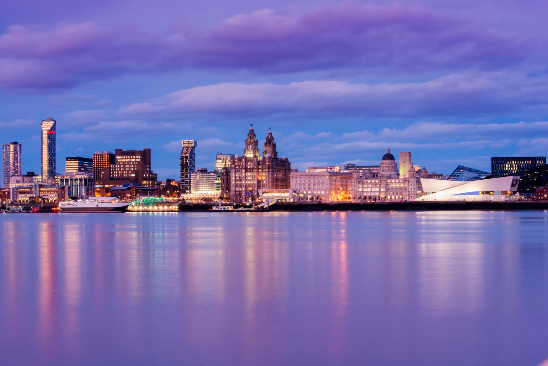 This is a horizontal, color photograph of Liverpool England at dusk. The docks are visible along the city skyline across the River Mersey.